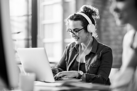hymyilevä nainen kirjoittaa tietokoneella kuulokkeet päässään